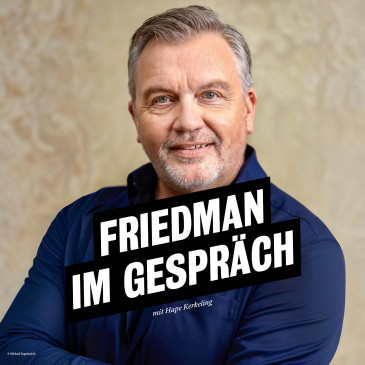 Hape Kerkeling trägt ein dunkelblaues Hemd, steht vor einer gelblichen Wand, hat die Arme verschränkt und schaut lächelnd direkt in die Kamera. 