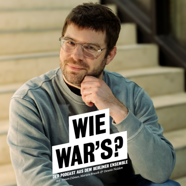 Portrait von Simon Dömer sitzt auf der Treppe im neuen Haus, er trägt einen grau-blauen Pullover und eine Brille, er fasst sich mit seiner linken Hand linksseitig ans Kinn.