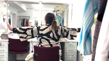 Timur sitzt mit dem Rücken zur Kamera vor einem großen querhängenden Spiegel im Maskenraum des Berliner Ensembles. Er trägt ein weites schwarz-weißes Oberteil und streckt die Arme wie Flügel zur Seite aus 