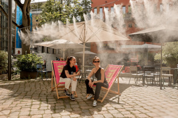 Zwei Personen sitzen bei Sonne auf Liegestühlen im Innenhof, darüber feiner Sprühregen aus zahlreichen Düsen