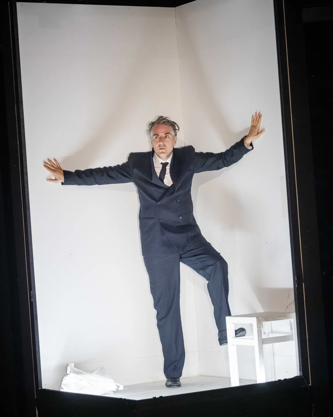 Jens Harzer im Bühnenbild. Er befindet sich in einem schmalen hohen und weißen Kasten, er trägt einen dunklen Anzug. 