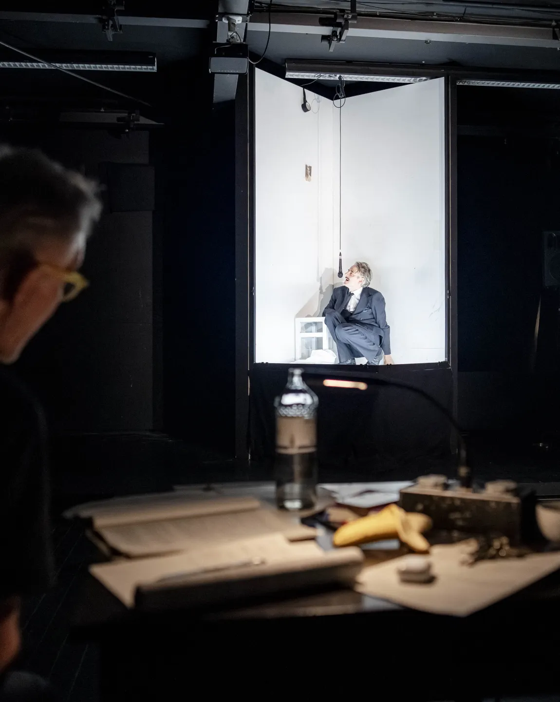 Jens Harzer kauert im weiß beleuchteten Bühnenbild, im vorderen, dunkleren, Bildteil ist Oliver Reese beobachtend im Profil zu erkennen. 