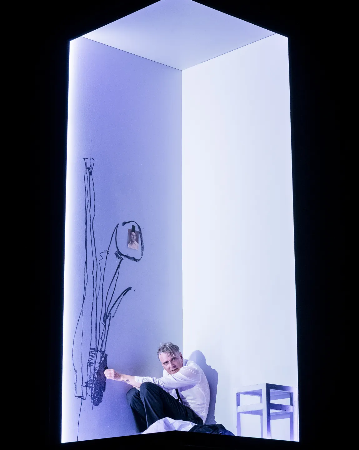 Schauspieler Jens Harzer kauert in einem beleuchteten Würfel auf dem Boden, neben ihm ist ein weißer Hocker, auf dem Boden liegen Klamotten, er skizziert an der Wand, daran klebt eine Fotografie.