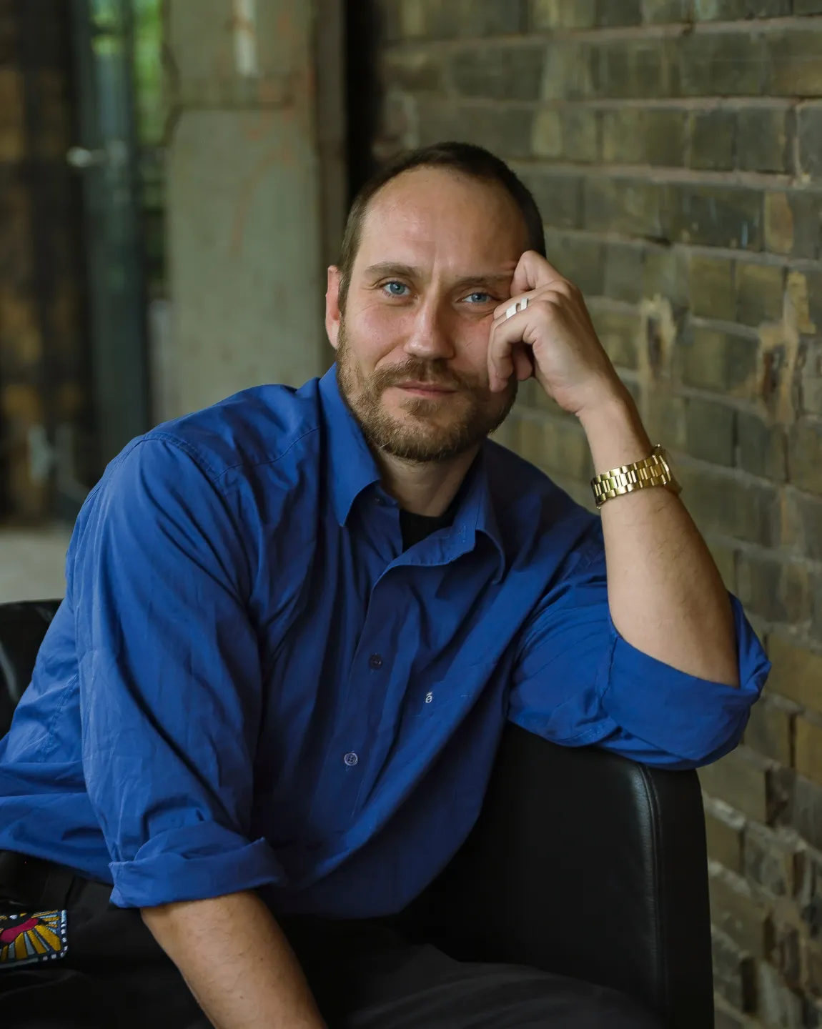 Portrait von Ensemblemitglied Sebastian Zimmler. Er sitzt in einem schwarzen Ledersessel vor einer Backsteinwand, trägt ein blaues Hemd, stützt sich auf seinen linken Arm auf und schaut direkt in die Kamera. 