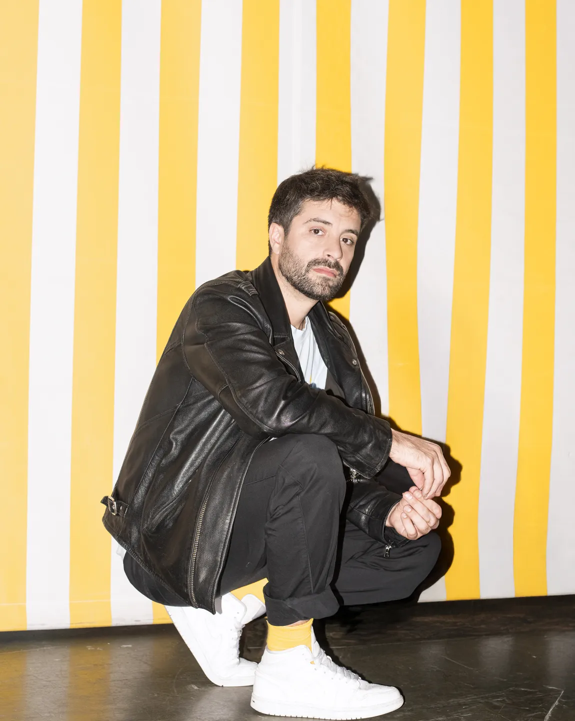 Regisseur Antú Romero Nunes trägt ein schwarzes Outfit samt Lederjacke, weiße Schuhe und hockt seitlich vor einer gelb-weiss-gestreiften Wand. 