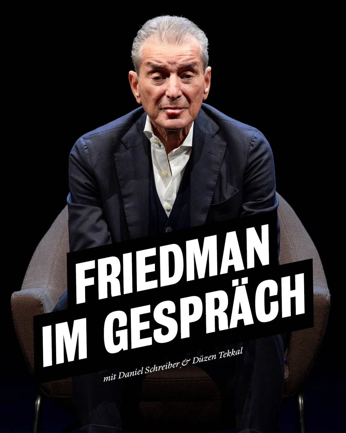 Portrait von Michel Friedman auf einem braunen Sessel sitzend, der Hintergrund ist dunkel. Er trägt einen dunklen Anzug mit weissem Kragen, lehnt sich etwas vor und schaut direkt in die Kamera. 