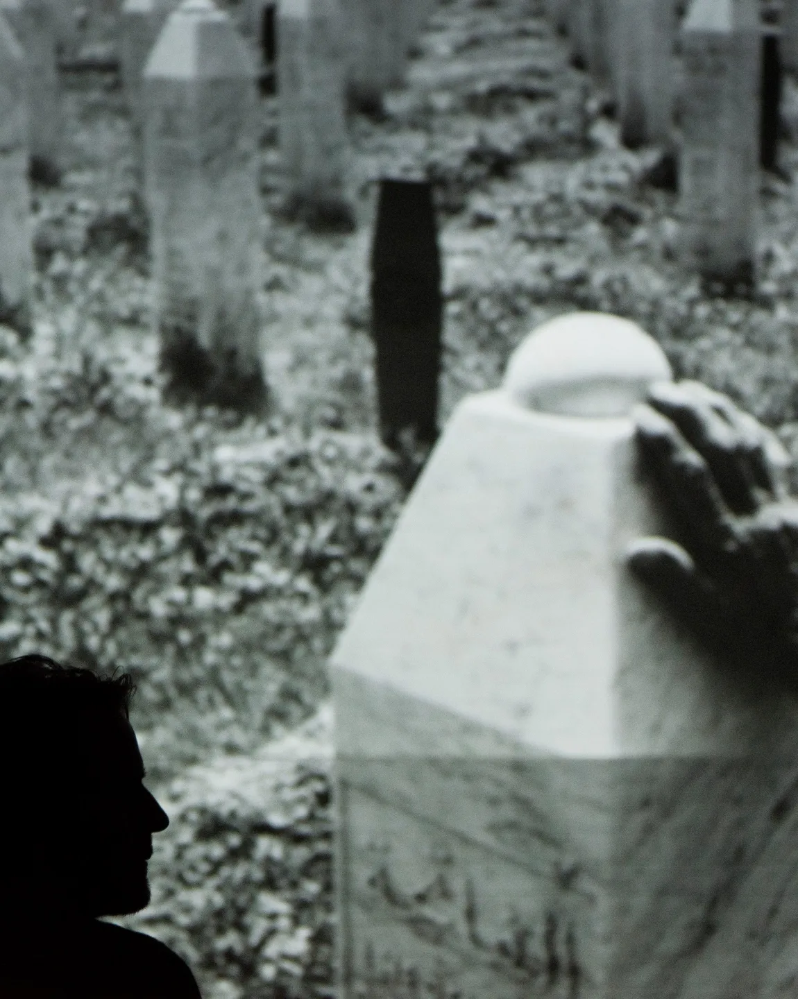 Inszenierungsfoto von Srebrenica, links im Bild die Umrisse von Jens Harzer vor einer Projektion eine Fotos im Hintergrund. Auf dem Foto sind Grabsteine in Form von Stelen zu erkennen sowie eine Hand, die einen dieser berührt. 