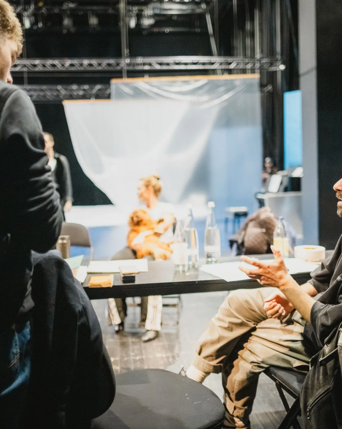 Regisseur Antú Romero Nunes sitzt am Regietisch und unterhält sich mit einem Kollegen, dessen schwarzer Hoodie mit der Aufschrift "Berliner Ensemble" der Kamera zugewandt ist. 
