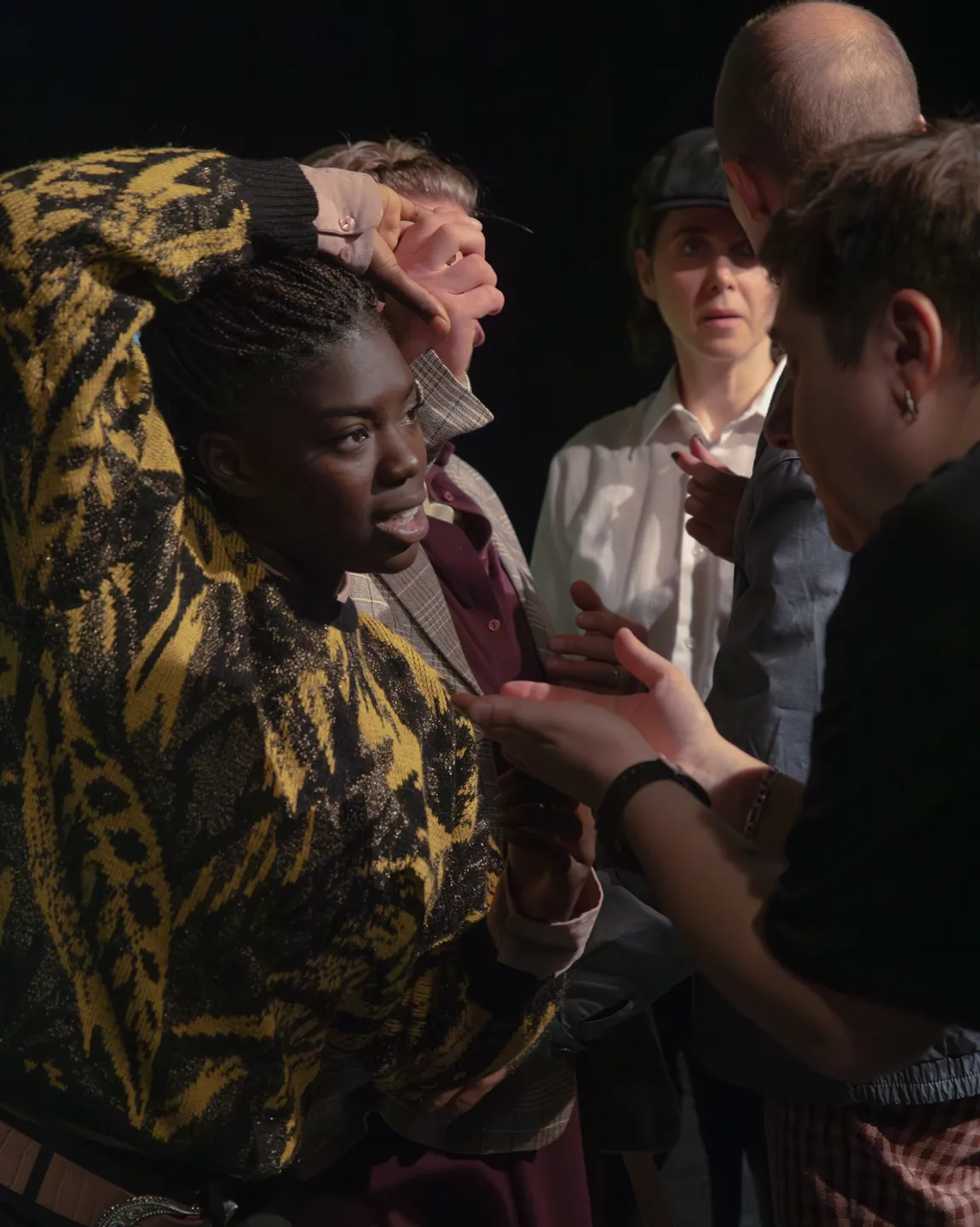 Joyce Sanhá und Stas Zhrykov im Gespräch. Sie hat ihren rechten Arm über ihren Kopf gelegt, im Hintergrund sind weitere Ensemblemitglieder zu erkennen. 