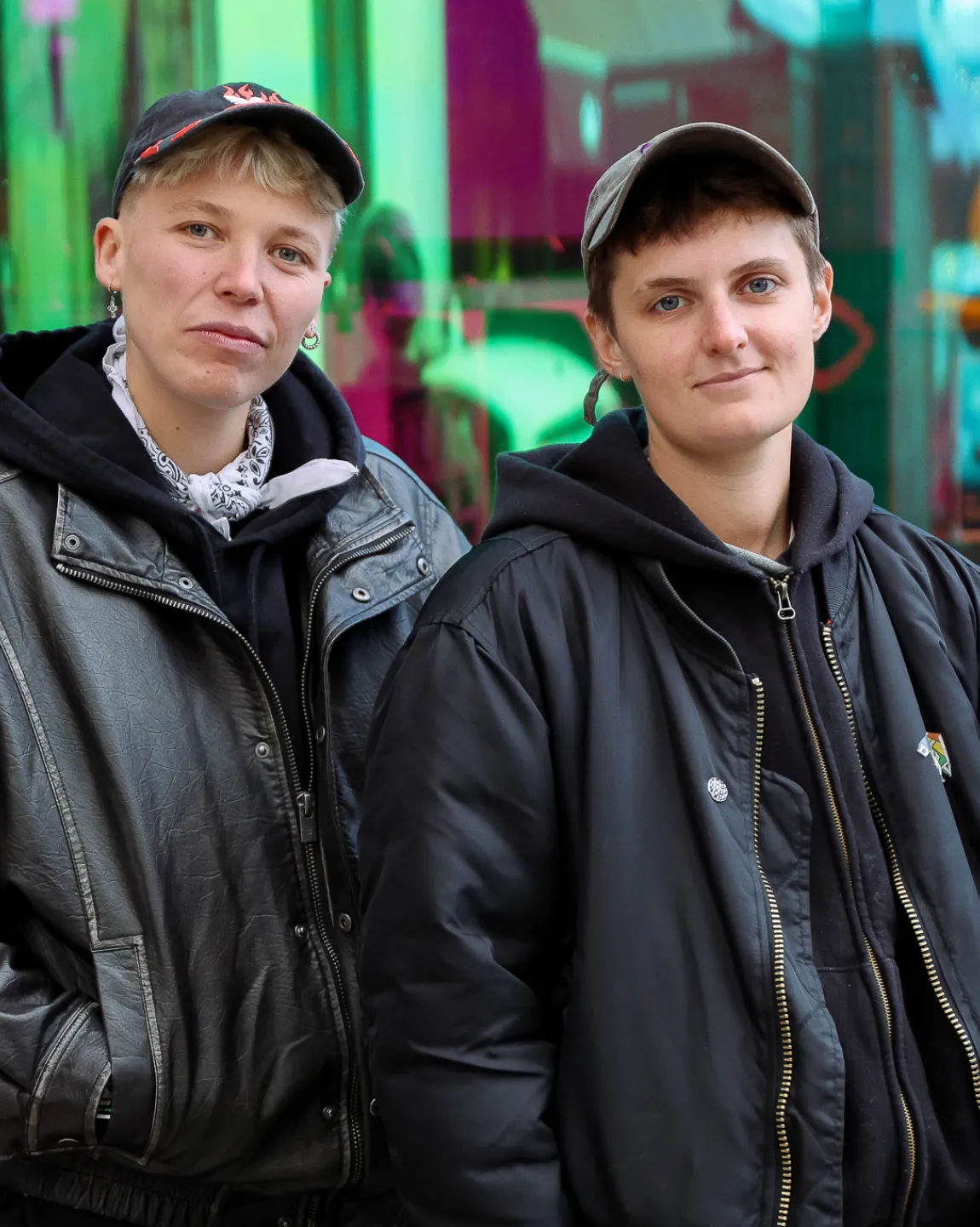 Jules Head und Leo Lorena Wyss stehen in schwarzen Bomberjacken und mit Caps vor der holografisch spiegelnden Glasfront des BE-Shops. Sie schauen ernst, stehen nah beieinander und schauen direkt in die Kamera. 