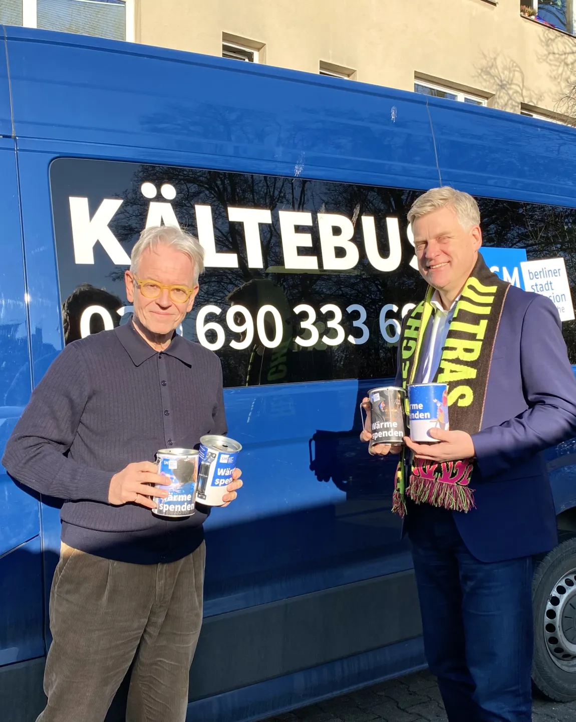 Oliver Reese übergibt dem Vorstand der Berliner Stadtmission die gesammelten Spenden. Sie stehen vor einem blauen Kältebus. Der Vorstand trägt einen Merchandiseschal des Berliner Ensembles. 