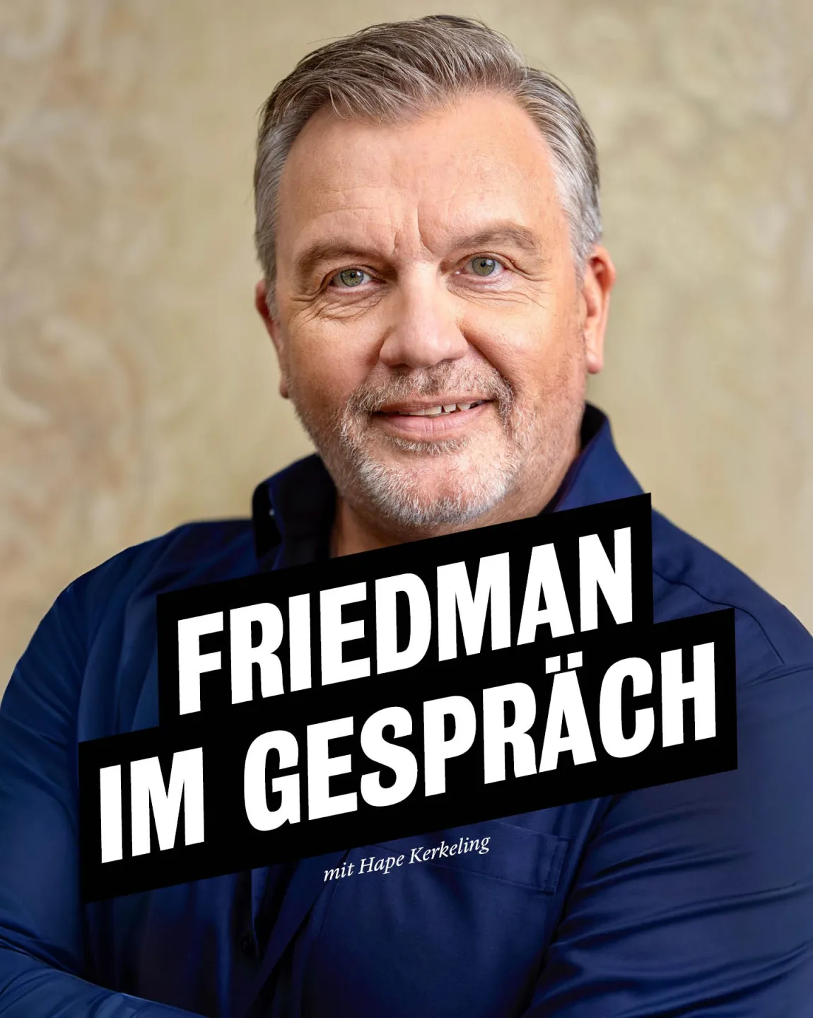 Hape Kerkeling trägt ein dunkelblaues Hemd, steht vor einer gelblichen Wand, hat die Arme verschränkt und schaut lächelnd direkt in die Kamera. 