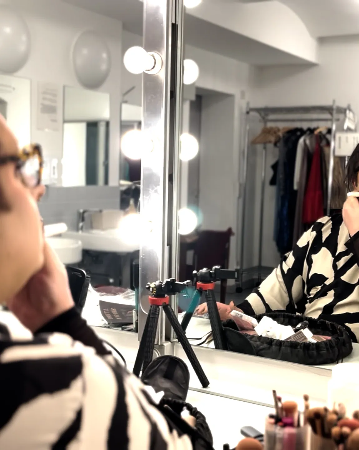 Der Creator Timur sitzt im Schminkraum vor einem großen Spiegel mit Glühbirnen. Links im Bild ist seine Schulter zu erkennen, das Foto zeigt aber sein Spiegelbild. Er ist bereits extravagant geschminkt und hält einen Pinsel in der Hand, mit dem er sich bepudert. 