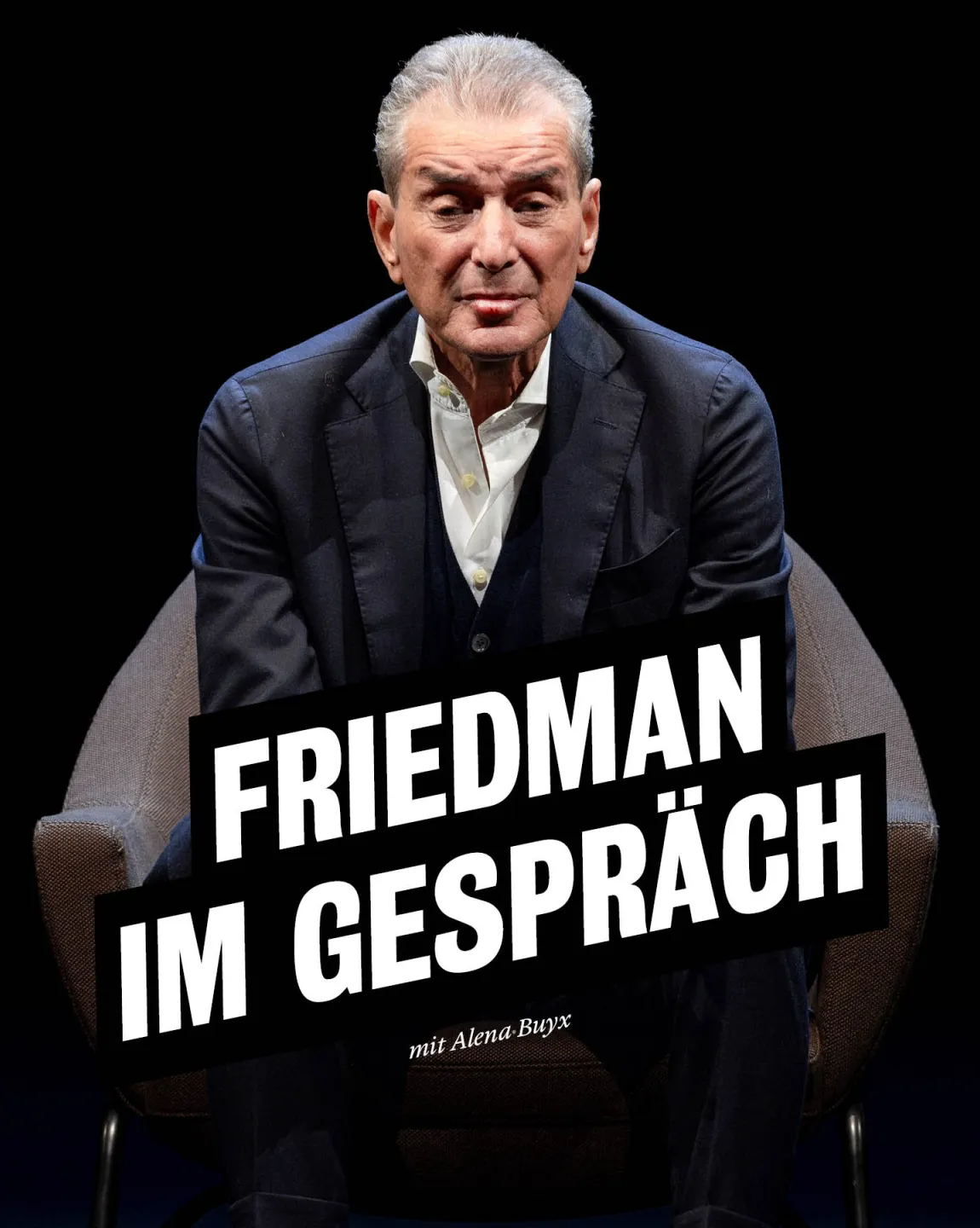 Michel Friedman trägt einen dunklen Anzug mit weißem Hemd, er sitzt auf einem dunklen Sessel vor dunklem Hintergrund. Er  lehnt sich leicht vor und schaut direkt in die Kamera. 