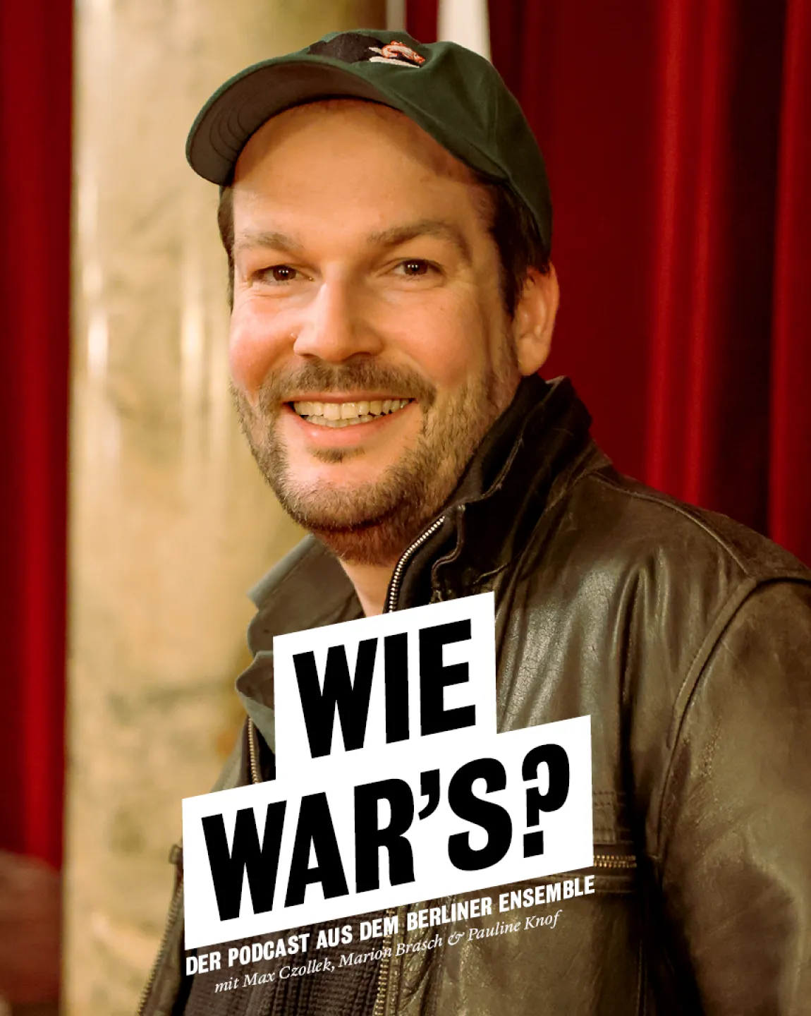 Portrait von Max Czollek in den Umgängen des Großen Hauses. Er lehnt an den Garderobentresen, hinter ihm sind rote SAmtvorhänge zu erkennen. Er trägt eine braune Lederjacke und eine grüne Cap, während er lachend direkt in die Kamera schaut. 