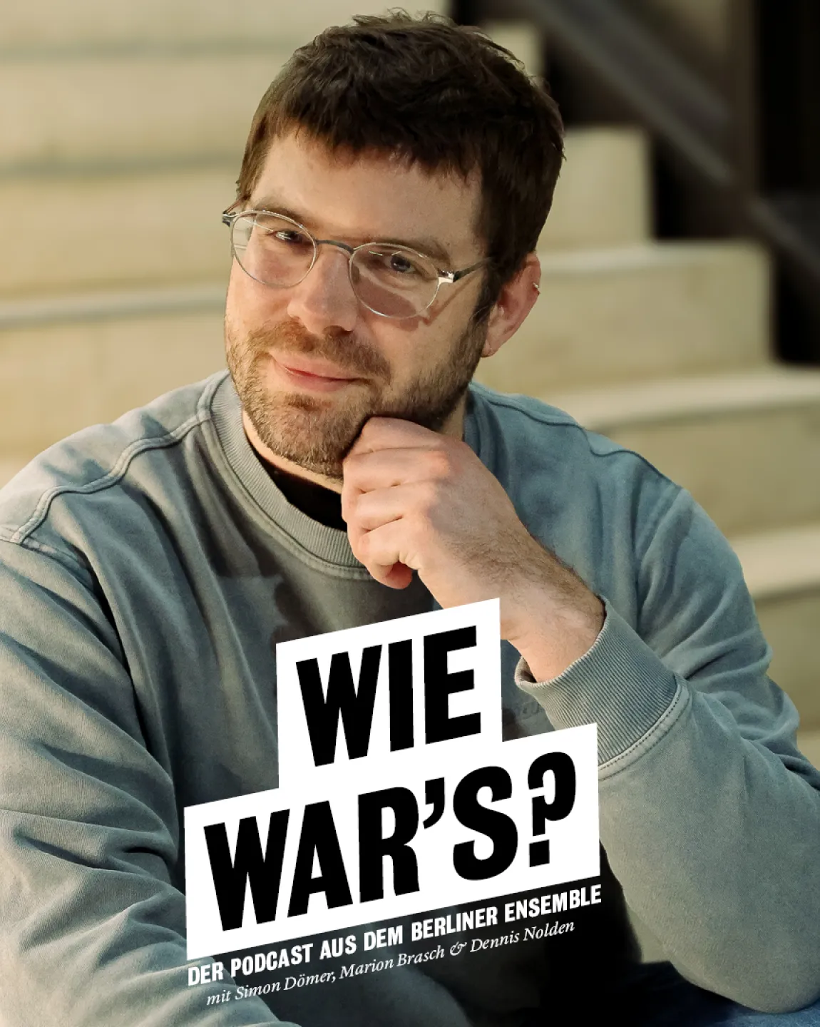 Portrait von Simon Dömer sitzt auf der Treppe im neuen Haus, er trägt einen grau-blauen Pullover und eine Brille, er fasst sich mit seiner linken Hand linksseitig ans Kinn.