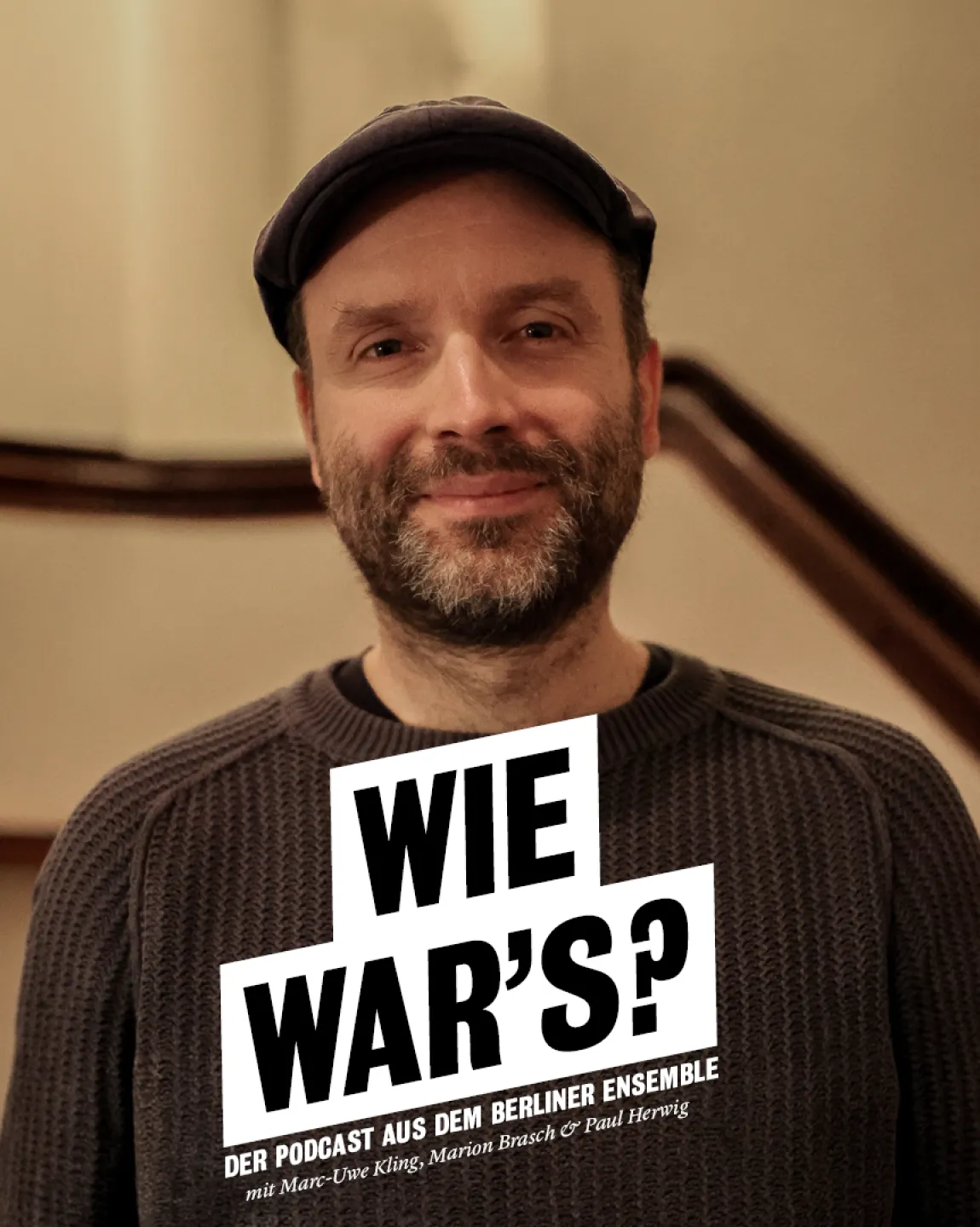 Portrait von Marc-Uwe Kling im Treppenaufgang zum ersten Rang im Großen Haus. Er trägt einen grau-braunen Pullover und eine Schiebermütze. Er lächelt direkt in die Kamera. 