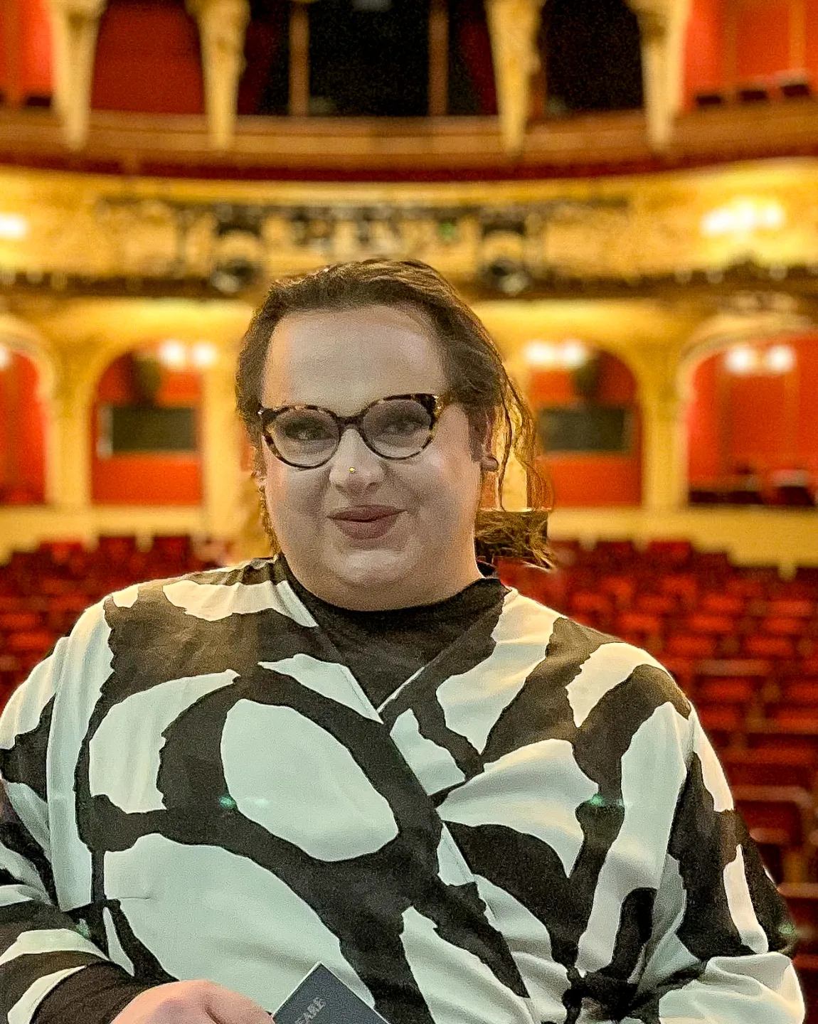 Timur steht auf der Bühne des Großen Hauses, im Hintergrund ist der  hell erleuchtete Zuschauerraum zu erkennen. Er schaut direkt in die Kamera. 