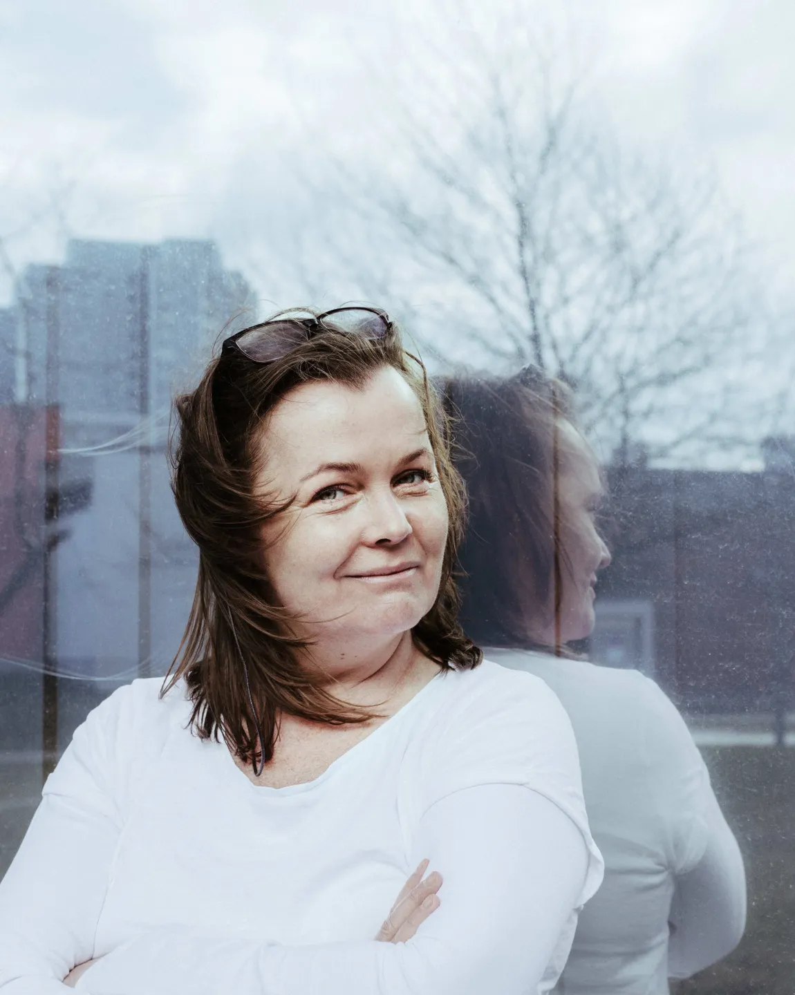 Portrait der Autorin Katja Oskamp vor einer spiegelnden Glasfläche. Es sind Hochhäuser zu erkennen. Sie trägt ein weißes Oberteil, verschränkt die Arme und schaut seitlich in die Kamera. 