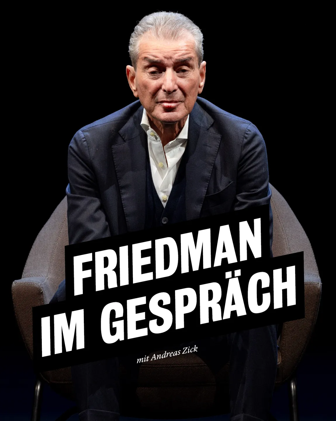 Michel Friedman sitzt auf einem dunklen Sessel vor dunklem Hintergrund. Er trägt einen dunklen Anzug über einem weißen Hemd und schaut, die Arme auf die Knie lehnend, direkt in die Kamera. 