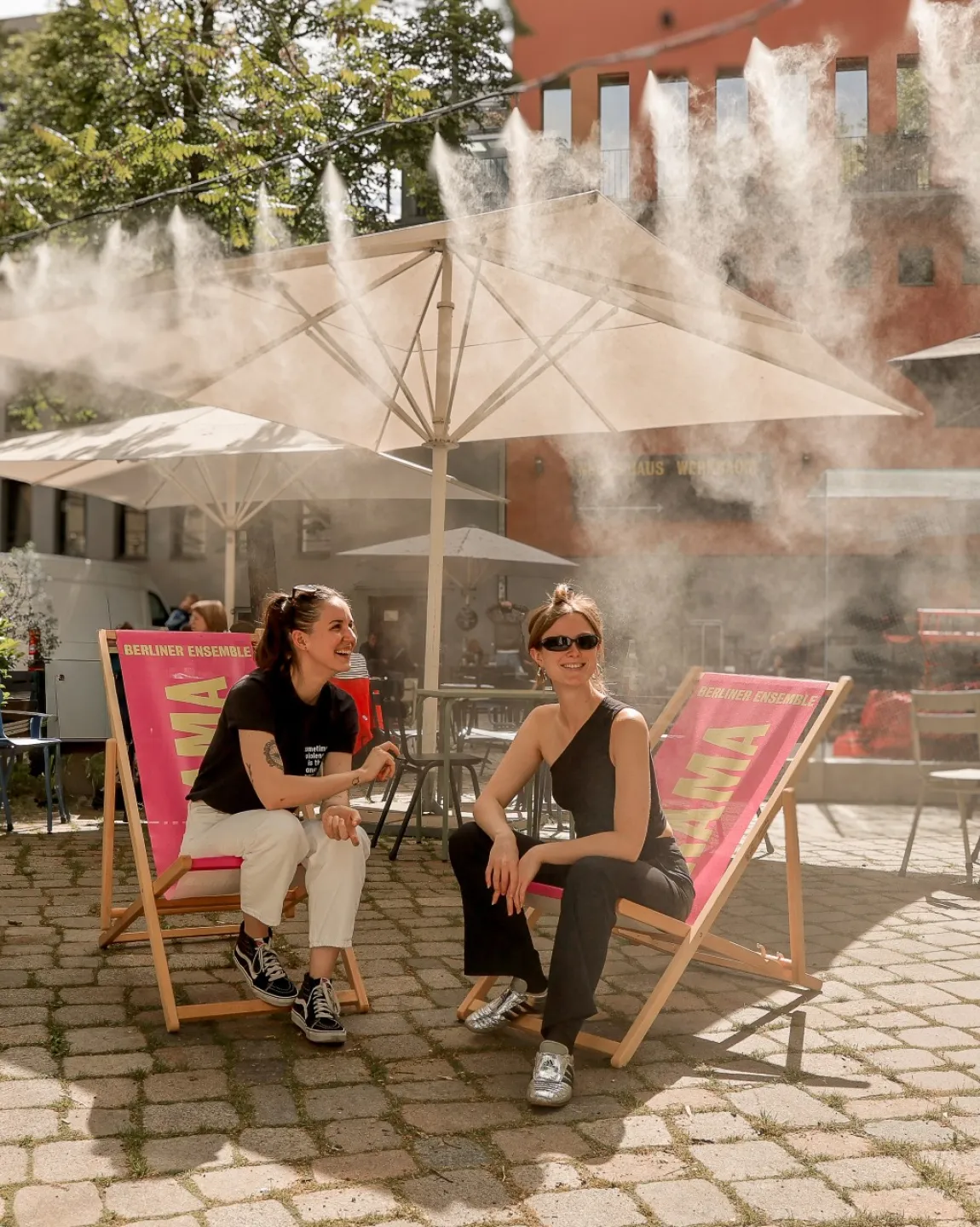Zwei Personen sitzen bei Sonne auf Liegestühlen im Innenhof, darüber feiner Sprühregen aus zahlreichen Düsen