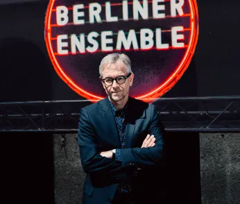 Oliver Reese steht mit verschränkten Armen vor dem Logo auf dem Dach des Berliner Ensembles. Das Logo leuchtet rot, der Hintergrund ist dunkel. 