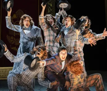 24)	Sieben Personen in karierten Anzügen stehen eng zusammen und strecken schwarze Zylinderhüte in die Luft.