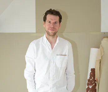 Ein Portrait des Künstlers Alex Wissel, er trägt ein weißes Hemd, schaut direkt in die Kamera und steht vor einer Wand. Im Hintergrund sind Tapetenrollen zu erkennen..