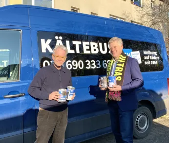 Oliver Reese übergibt dem Vorstand der Berliner Stadtmission die gesammelten Spenden. Sie stehen vor einem blauen Kältebus. Der Vorstand trägt einen Merchandiseschal des Berliner Ensembles. 