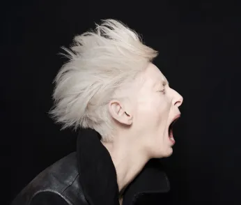 Aufnahme von Tilda Swinton im Seitenprofil. Der Hintergrund ist schwarz. Sie trägt eine schwarze Lederjacke mit Kragen, hat kurze platinblonde Haare und schreit.