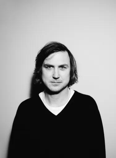 Schwarz-weiss-Aufnahme von Schauspieler Lars Eidinger vor hellem Studiohintergrund in dunklem Pullover, er schaut frontal in die Kamera