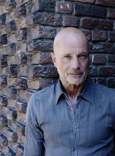Schauspieler Christian Berkel in blaugrauem Hemd vor einer Backsteinmauer, schaut in die Kamera. 