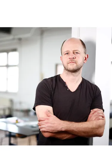 Jan Hendrik Neidert lehnt mit verschränkten Armen an einem weißen Pfeiler. Im Hintergrund ist unscharf eine Art Großraumbüro zu erkennen. Er trägt ein braunes Shirt.