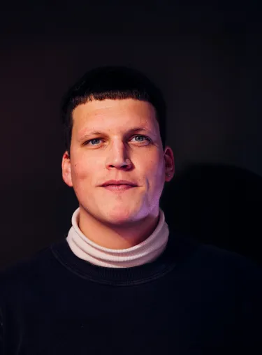 Potraitaufnahme von August Modersohn in schwarzem Oberteil mit weißem Kragen vor schwarzem Hintergrund, er lächelt und schaut in die Kamera.