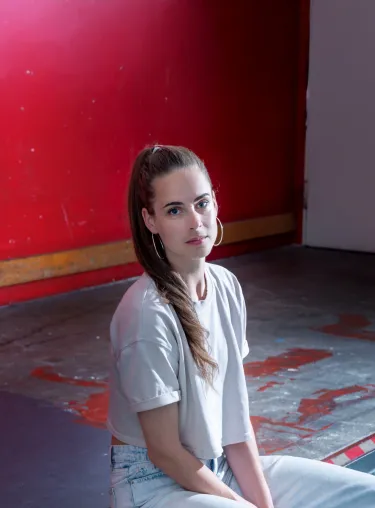 Schauspielerin Tabitha Frehner sitzt in hellem Shirt und Hose an der Kante vor einer roten Wand.