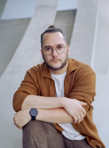 Portraitfoto von Dramaturg Daniel Grünauer, er sitzt, lehnt seine beiden Arme auf das rechte Knie und schaut direkt in die Kamera. 