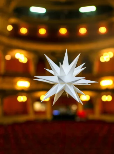 In der Mitte des Bildes ist ein weisser Weihnachtsstern zu erkennen, im Hintergrund leuchtet der Zuschauerraum des Berliner Ensembles Rot und Gold ausgeleuchtet. 