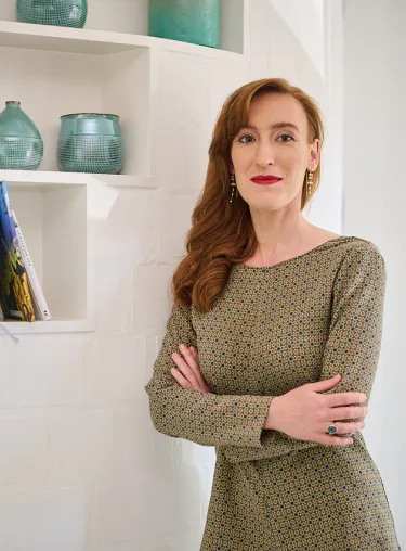 Nora Bossing in gemustertem Kleid mit verschränkten Armen vor einem weißen Wandregal mit Büchern und türkisen Vasen.