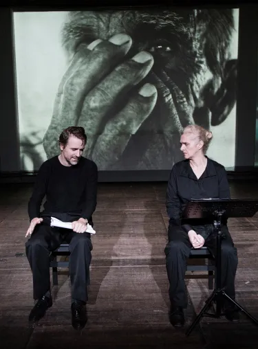 Inszenierungsfoto von Srebrenica, links im Bild Jens Harzer, rechts Vernesa Berbo, die zum Publikum gewandt auf der Bühne sitzen. Mittig im Hintergrund eine schwarz-weiß Projektion einer Nahaufnahme eines Mannes, der sich die Hände vors Gesicht hält, links der Ausschnitt einer Aufnahme eines brennenden Hauses, rechts ein weinendes Kind. Rechts und links auf der Bühne liegen kleine Haufen Holzscheite.