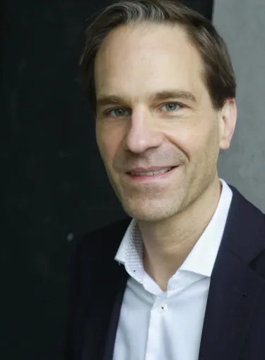 Volker Kitz in weißem Hemd und dunklem Anzug vor dunkler Wand, schaut in die Kamera.