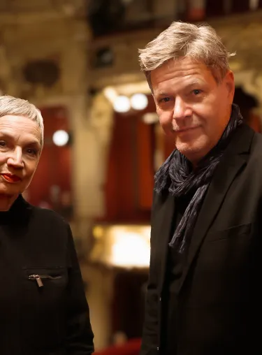 Robert Habeck und Marion Brasch stehen in einer Loge, im Hintergrund ist der Zuschauerraum zu erkennen. Sie tragen beide dunkle Kleidung und schauen direkt in die Kamera.
