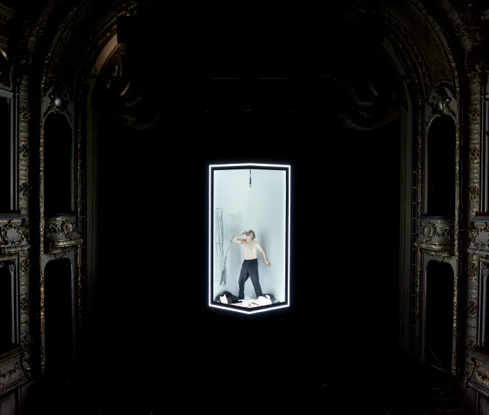 09)	Oberkörperfreier Mann  steht mit einem Mikro in der Hand in einem beleuchteten, rechteckigen Kasten auf einer dunklen Bühne. Es sind Teile des neobarocken Portals und der Logen des Theaters zu sehen. 