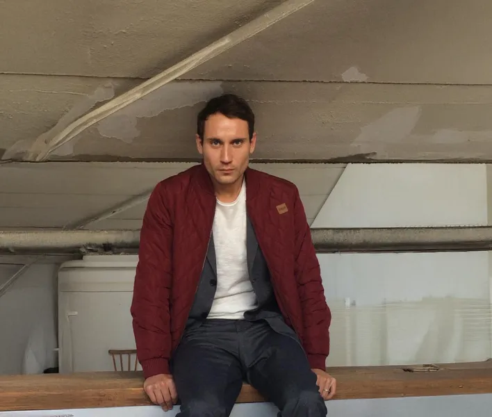 Schauspieler Sebastian Zimmler sitzt in roter Jacke mit weißem Shirt und dunkler Hose vor grauer Wand auf einem Holztresen.