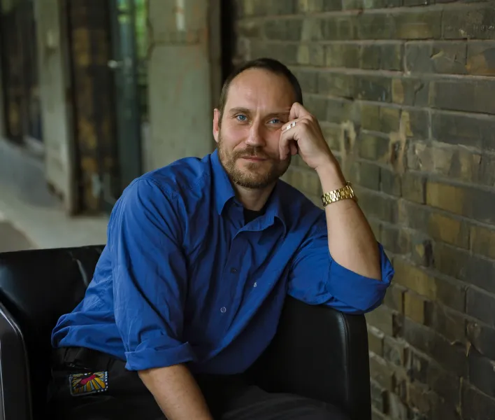 Portrait von Ensemblemitglied Sebastian Zimmler. Er sitzt in einem schwarzen Ledersessel vor einer Backsteinwand, trägt ein blaues Hemd, stützt sich auf seinen linken Arm auf und schaut direkt in die Kamera. 