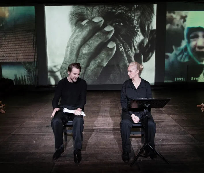 Inszenierungsfoto von Srebrenica, links im Bild Jens Harzer, rechts Vernesa Berbo, die zum Publikum gewandt auf der Bühne sitzen. Mittig im Hintergrund eine schwarz-weiß Projektion einer Nahaufnahme eines Mannes, der sich die Hände vors Gesicht hält, links der Ausschnitt einer Aufnahme eines brennenden Hauses, rechts ein weinendes Kind. Rechts und links auf der Bühne liegen kleine Haufen Holzscheite.