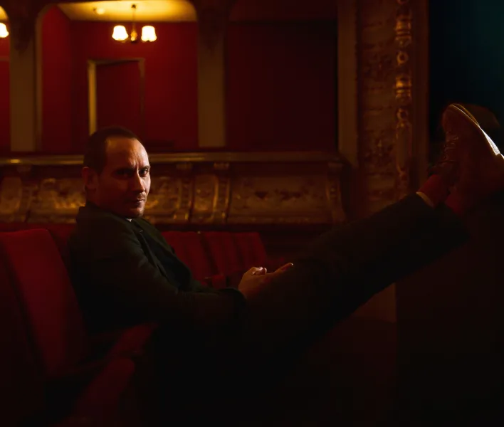 Foto von Sebastian Zimmler im Zuschauerraum des Großen Hauses. Im Hintergrund sind die Parkettlogen zu erkennen, das Bild ist rötlich gefärbt. Sebastian Zimmler sitzt auf einem Samtstuhl, legt die Füße auf den Vordersitz und schaut in die Kamera.