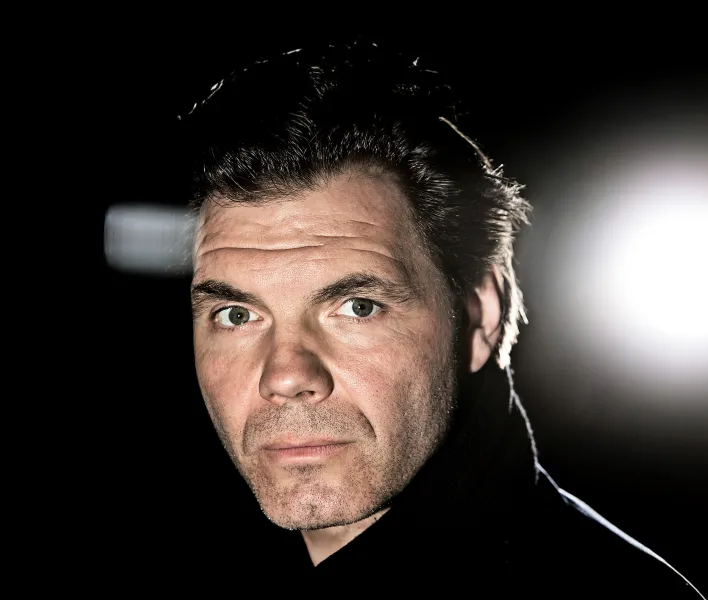 Ein Portrait von max Hopp. Er schaut über die linke Schulter direkt in die Kamera. Das Foto wurde mit Blitz aufgenommen, im Hintergrund ist ein leuchtender Scheinwerfer zu erkennen. 